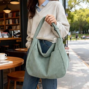 Puede incluir: Un bolso de hombro grande de pana verde salvia con forma rectangular y dos bolsillos delanteros. El bolso tiene una correa larga y un peque&ntilde;o dije de perro. La persona lleva un su&eacute;ter gris claro y vaqueros azules.