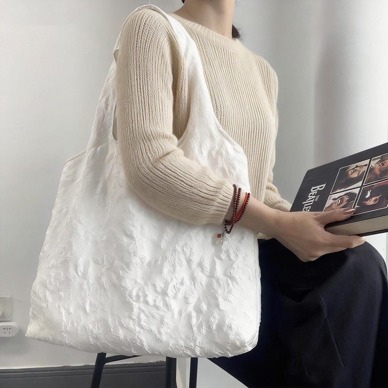 Pu&ograve; includere: Una grande borsa tote bianca con un motivo floreale strutturato. La borsa ha un'ampia tracolla ed &egrave; realizzata in un materiale morbido, forse simile al lino. Una persona indossa la borsa e indossa un maglione color crema.