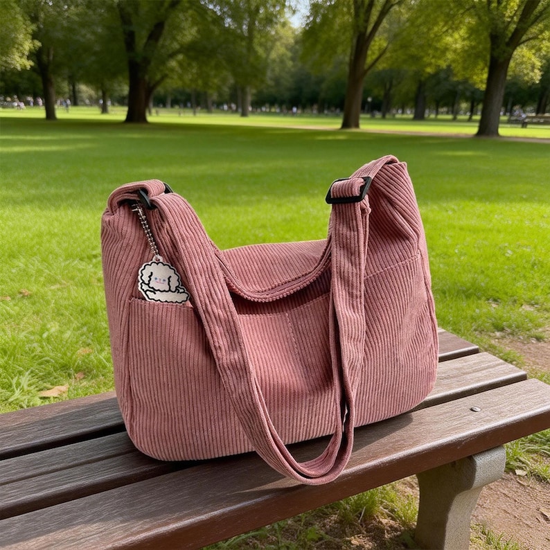 Puede incluir: Bolso de hombro de pana rosa polvoriento con solapa y correa ajustable. Tiene un peque&ntilde;o bolsillo delantero y un charm decorativo. El bolso est&aacute; sobre un banco de madera en un parque.