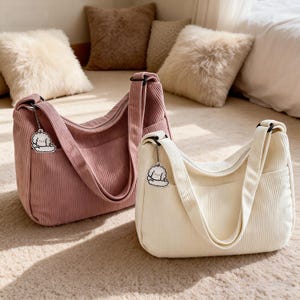 Puede incluir: Dos bolsos de hombro de pana en la imagen. Uno es de color rosa empolvado y el otro blanco roto. Cada bolso tiene una correa a juego, un bolsillo frontal y un cierre de cremallera. Un peque&ntilde;o dije de una nube con un perro est&aacute; adjunto a cada bolso.