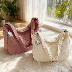 Puede incluir: Dos bolsos de hombro de pana en rosa y crema. Cada bolso tiene una correa larga, un bolsillo delantero y un cierre de cremallera. Un peque&ntilde;o dije de un perro de dibujos animados cuelga de cada bolso.