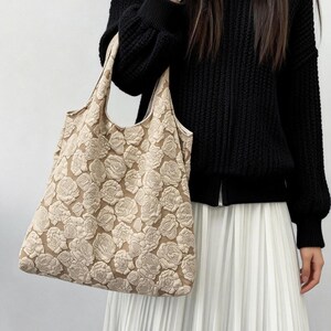 Pu&ograve; includere: Borsa tote beige con un motivo floreale ripetuto. La borsa &egrave; tenuta da una persona che indossa un maglione nero e una gonna plissettata bianca. Il design della borsa presenta rose strutturate in rilievo.