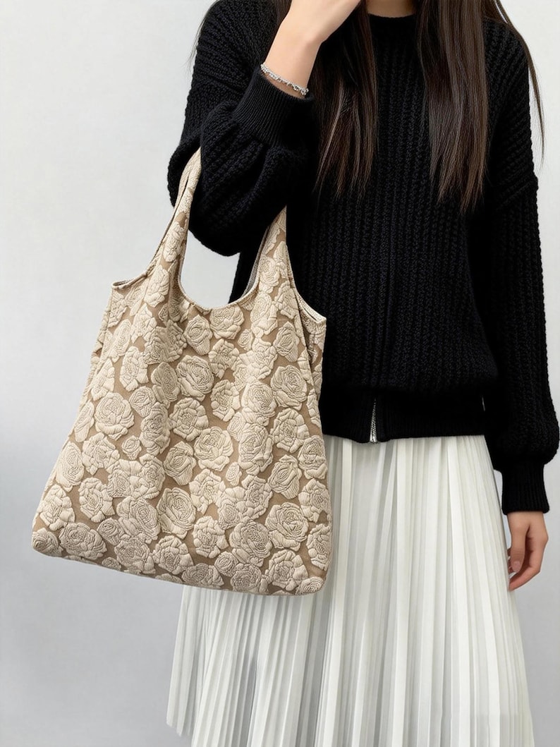 Pu&ograve; includere: Borsa tote beige con un motivo floreale ripetuto. La borsa &egrave; tenuta da una persona che indossa un maglione nero e una gonna plissettata bianca. Il design della borsa presenta rose strutturate in rilievo.