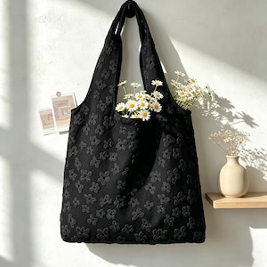 May include: A black tote bag featuring an all-over textured floral pattern. The bag has two shoulder straps and is filled with white daisies with yellow centers. The fabric has a raised, three-dimensional quality to the flower shapes.