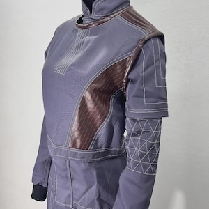 May include: A gray jumpsuit with brown leather-like accents on the shoulders and chest. The jumpsuit features white stitching details and a high collar. The sleeves have geometric patterns near the cuffs. The mannequin is wearing a black glove.