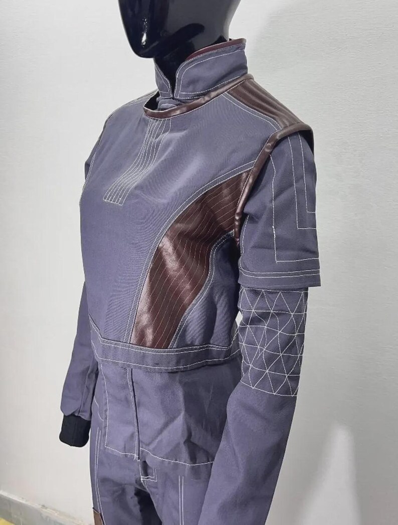 May include: A gray jumpsuit with brown leather-like accents on the shoulders and chest. The jumpsuit features white stitching details and a high collar. The sleeves have geometric patterns near the cuffs. The mannequin is wearing a black glove.