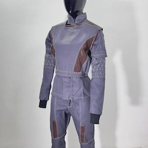 May include: A full-body jumpsuit in muted blue-gray with brown accents on the shoulders and knees. The suit features white stitching details and black cuffs. The mannequin's head is black and glossy.