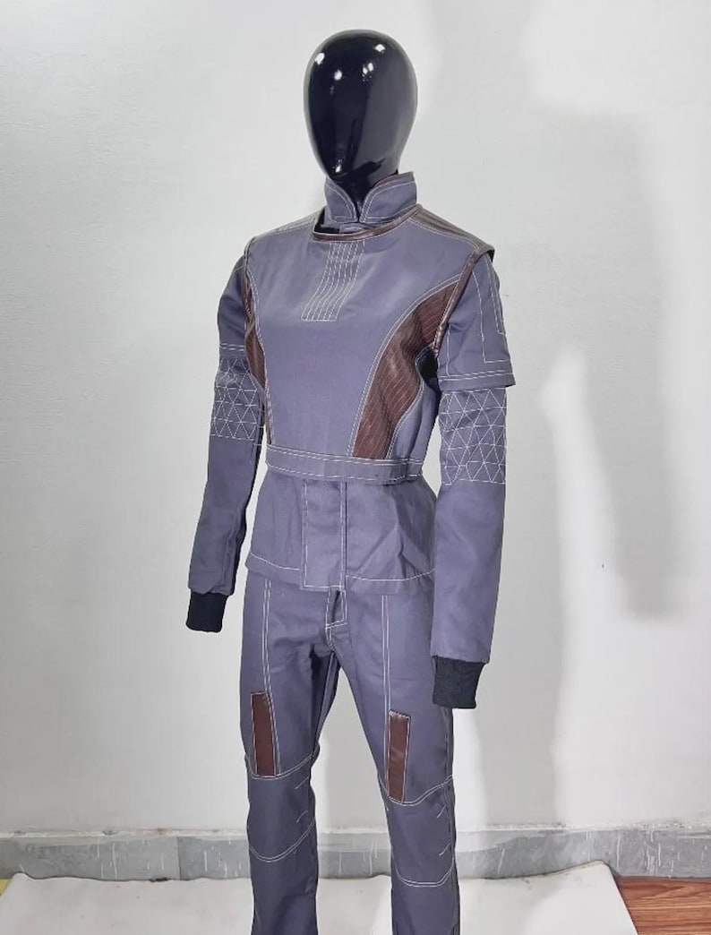 May include: A full-body jumpsuit in muted blue-gray with brown accents on the shoulders and knees. The suit features white stitching details and black cuffs. The mannequin's head is black and glossy.