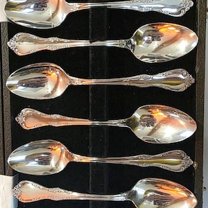 May include: A set of six silver spoons presented in a black velvet-lined box. Each spoon features a rounded bowl and decorative, floral detailing on the handle. The spoons are arranged neatly, reflecting light.