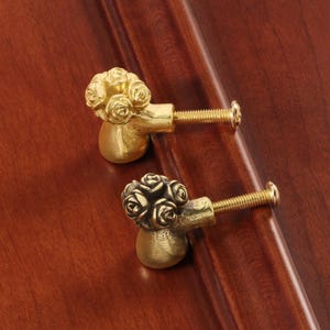 Könnte beinhalten: Zwei Messing-Schrankknöpfe mit Rosenstrauß-Design. Die goldfarbenen Knöpfe haben eine Gewindeschraube zur Befestigung. Sie sind auf einer dunkelbraunen Holzoberfläche montiert.