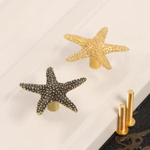 Könnte beinhalten: Zwei sternförmige Schrankknöpfe, einer goldfarben und einer dunkelgrau mit goldenen Akzenten, liegen auf einer weißen Oberfläche. Zwei goldfarbene Schrauben sind ebenfalls sichtbar. Diese dekorativen Knöpfe verleihen Möbeln einen maritimen Touch.