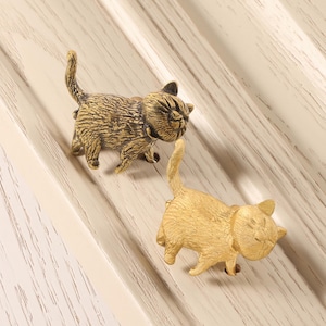 Könnte beinhalten: Zwei messingfarbene Katzen-Schrankknöpfe. Ein Knopf hat eine strukturierte, detaillierte Oberfläche, während der andere glatt ist. Beide Knöpfe befinden sich in einer Laufposition, wobei ihre Schwänze angehoben sind. Die Knöpfe sind auf einer hellen Holzoberfläche montiert.