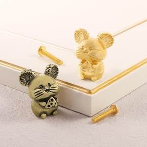 Könnte beinhalten: Zwei dekorative Schrankknöpfe in Form von Mäusen, die Käse halten. Die Knöpfe sind goldfarben und haben ein detailliertes, strukturiertes Design. Zwei goldene Schrauben sind ebenfalls sichtbar. Diese Knöpfe eignen sich perfekt, um Möbeln eine skurrile Note zu verleihen.