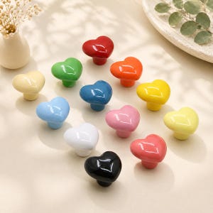 Könnte beinhalten: Eine Sammlung herzförmiger Schrankknöpfe in verschiedenen Farben, darunter Rot, Grün, Orange, Blau, Rosa, Gelb, Weiß und Schwarz. Diese dekorativen Knöpfe sind aus Keramik gefertigt und haben eine glänzende Oberfläche.