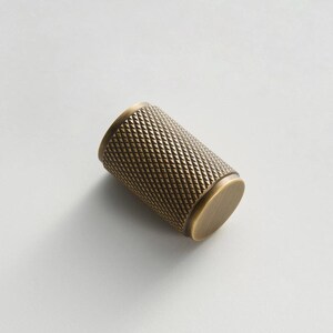 May include: A cylindrical cabinet knob with a diamond pattern texture in a brass finish. The knob has a flat, circular end on each side. The hardware is designed for use on drawers and cabinets.