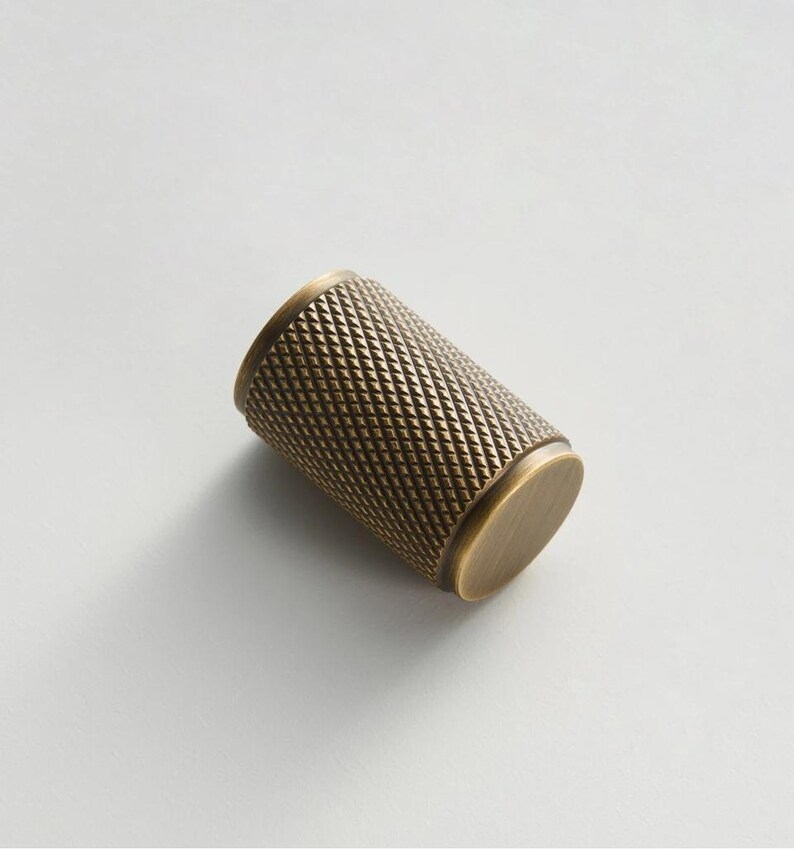 May include: A cylindrical cabinet knob with a diamond pattern texture in a brass finish. The knob has a flat, circular end on each side. The hardware is designed for use on drawers and cabinets.
