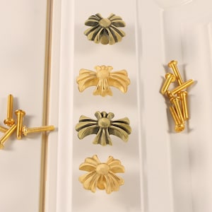 Könnte beinhalten: Vier dekorative Schrankknöpfe mit einem blütenartigen Design in Antikgold- und Bronzetönen. Mehrere goldene Schrauben sind ebenfalls sichtbar. Die Knöpfe sind an einer weißen Schranktür montiert, was auf ein Wohndekorations-Thema hindeutet.