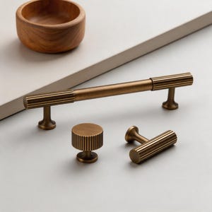 May include: A set of antique brass cabinet hardware, including a long handle, a knob, and a smaller handle. The hardware features a ribbed design and a warm, metallic finish. A wooden bowl is in the background.