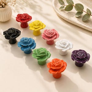 May include: A collection of colorful ceramic rose-shaped cabinet knobs. The knobs are in various colors, including red, yellow, black, blue, pink, white, green, orange, and purple. Each knob has a detailed rose design.
