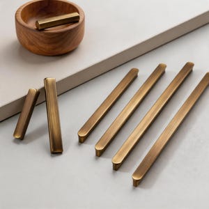 May include: A selection of antique brass cabinet pulls and a small brass bar in a wooden bowl. The pulls are rectangular with rounded ends. The bowl is round and made of wood. The background is a neutral colour.