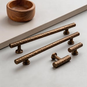 May include: A set of bronze-colored cabinet pulls and knobs with a hammered texture. The hardware includes two long pulls, a pair of smaller knobs, and a single T-shaped knob. A wooden bowl sits in the background.
