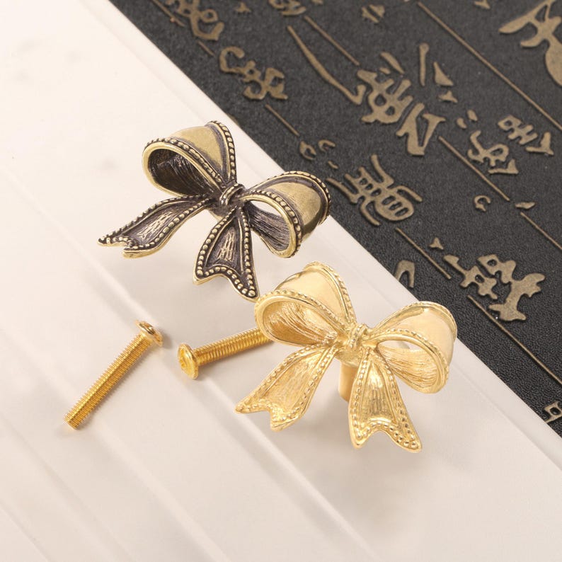 May include: Two decorative bow-shaped cabinet pulls in antique gold and bronze finishes. Each pull features a detailed bow design with a screw for installation. The pulls are displayed on a white surface with a dark background.