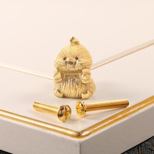 Könnte beinhalten: Goldfarbenes Möbelbeschlagsset mit einem dekorativen knauf in Kükenform und zwei passenden Schrauben. Der Kükenknauf hat eine strukturierte Oberfläche. Die Schrauben haben einen Kreuzschlitz.