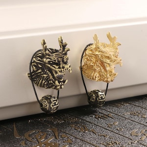 Könnte beinhalten: Zwei dekorative Schubladengriffe mit Drachenkopf. Einer ist bronzefarben, der andere goldfarben. Jeder Drachenkopf ist an einer schwarzen Schnur und einem dekorativen Ball befestigt. Die Schubladengriffe sind auf einer weißen Oberfläche montiert.