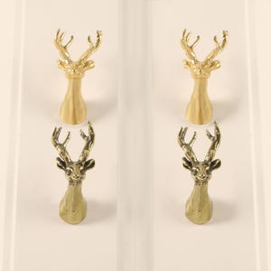 May include: Four decorative cabinet knobs with a deer head design. Two are gold-colored, and two are bronze-colored. The knobs are mounted on a white surface, showcasing their detailed antler and face features. These knobs add a rustic touch to any furniture.