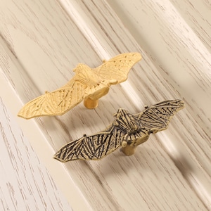 以下が含まれることがあります： 2つの装飾的なコウモリ型のキャビネットプル。1つは明るい金色、もう1つは濃いブロンズ色です。コウモリの翼は広げられており、プルは明るい色の木製の表面に取り付けられています。家具やキャビネットのハードウェアに適しています。