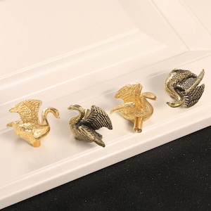 May include: Four swan-shaped cabinet knobs in gold and bronze tones. The metal knobs are displayed on a white surface. Each swan features detailed wing and feather designs, adding a touch of elegance to any cabinet.