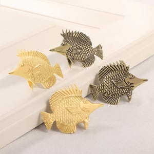 Könnte beinhalten: Vier dekorative Schrankknöpfe in Fischform in Gold- und Bronzetönen. Die Knöpfe sind aus Metall gefertigt und weisen detaillierte Schuppen, Flossen und eine spitze Schnauze auf. Die Knöpfe sind auf einer weißen Oberfläche angeordnet.