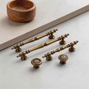 Op de afbeelding: Een assortiment antieke messing kastbeslagen, waaronder drie handgrepen en twee ronde knoppen. De handgrepen hebben een gedetailleerd, sierlijk ontwerp en de knoppen hebben een zonnestraalpatroon. Een houten kom staat op de achtergrond.