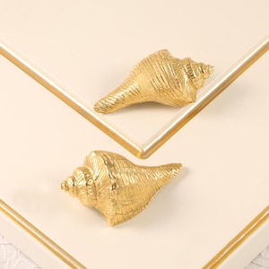 Op de afbeelding: Twee goudkleurige schelpenkastgrepen. De grepen hebben de vorm van schelpen met gedetailleerde texturen. Ze zijn geplaatst op een wit oppervlak met een gouden rand, wat een kust- of nautisch thema creëert.