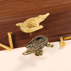 May include: Two decorative alligator-shaped cabinet knobs in gold and bronze tones. The knobs are detailed with textured scales and are shown with gold-colored screws. The knobs are likely for furniture or decorative use.