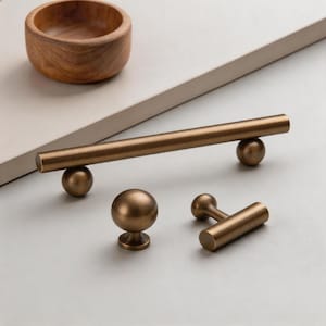Könnte beinhalten: Eine Sammlung von bronzefarbenen Schrankbeschlägen, darunter ein langer Stangengriff mit Kugelakzenten, ein runder Knopf und ein T-förmiger Griff. Im Hintergrund befindet sich eine kleine Holzschale. Die Hardware hat ein mattes Finish.