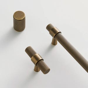 May include: A collection of brass cabinet hardware, including a cylindrical knob and two T-bar pulls. The hardware features a textured, cross-hatched pattern and a warm, antique brass finish. The word "NURLIN" is printed on a blue rectangle.