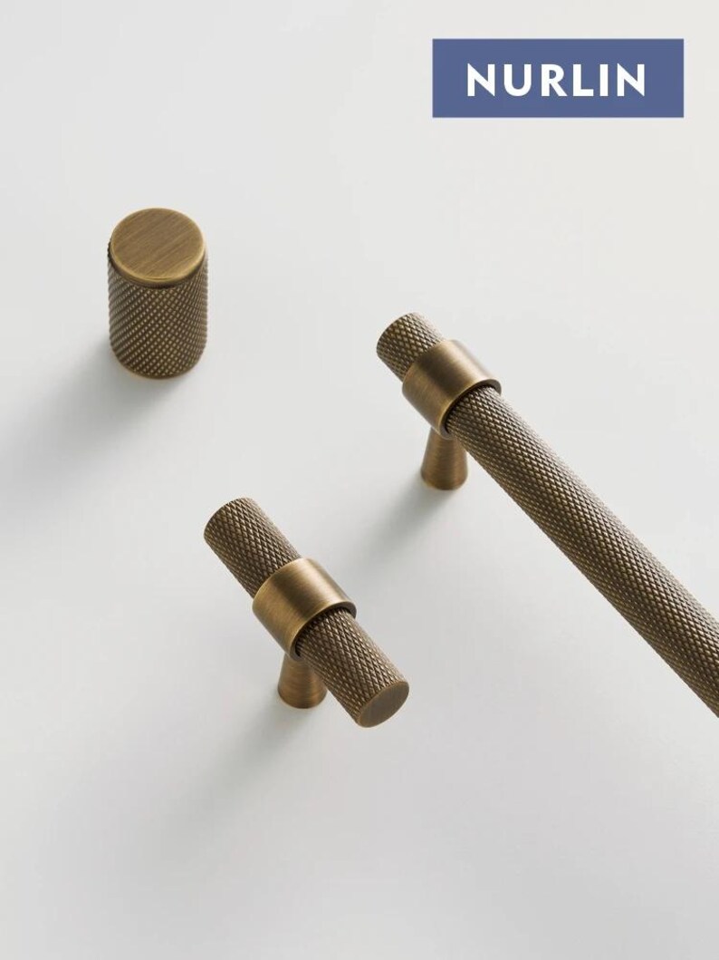 May include: A collection of brass cabinet hardware, including a cylindrical knob and two T-bar pulls. The hardware features a textured, cross-hatched pattern and a warm, antique brass finish. The word "NURLIN" is printed on a blue rectangle.