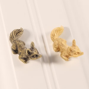 Könnte beinhalten: Zwei Eichhörnchen-Figuren. Eine ist dunkelbronze, die andere hellgoldfarben. Beide befinden sich in einer gekrümmten Position mit gewölbten Schwänzen. Die Figuren befinden sich auf einer weißen Oberfläche.