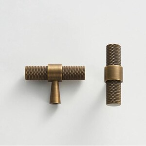 May include: Two antique brass cabinet pulls. One is horizontal with a T-shaped base, and the other is vertical. Both have a textured, diamond-patterned grip and a smooth, cylindrical band.