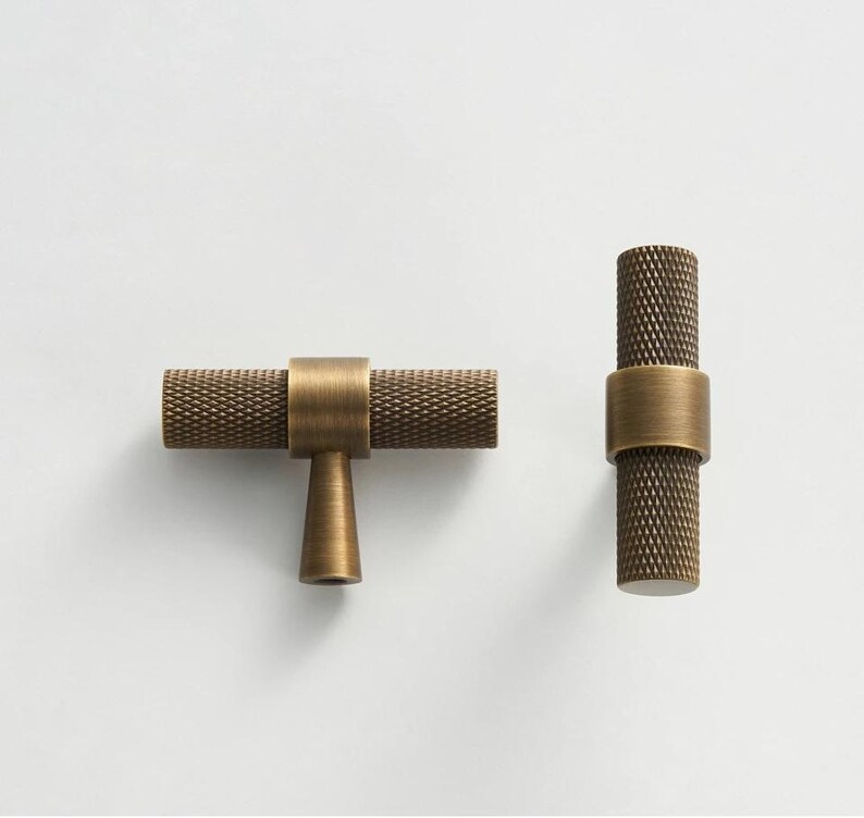 May include: Two antique brass cabinet pulls. One is horizontal with a T-shaped base, and the other is vertical. Both have a textured, diamond-patterned grip and a smooth, cylindrical band.
