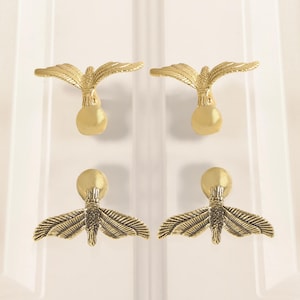 May include: Four gold-colored cabinet pulls. Two pulls feature an eagle design with outstretched wings, and two have a moth-like design with detailed wing patterns. The pulls are mounted on a white surface.