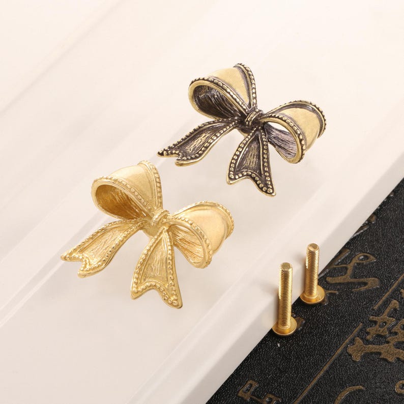 May include: Two bow-shaped cabinet pulls in antique gold and bronze tones. The decorative hardware features detailed bow designs with textured surfaces. Two gold-coloured screws are also visible, suggesting easy installation. These pulls are ideal for adding a touch of elegance to furniture.