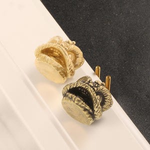 Könnte beinhalten: Zwei Messing-Schrankknöpfe mit einem Widderkopf-Design. Die Knöpfe sind goldfarben und haben eine detaillierte, strukturierte Oberfläche. Jeder Knopf hat zwei Schraubpfosten zur Befestigung. Die Knöpfe werden auf einem weißen und schwarzen Hintergrund präsentiert.