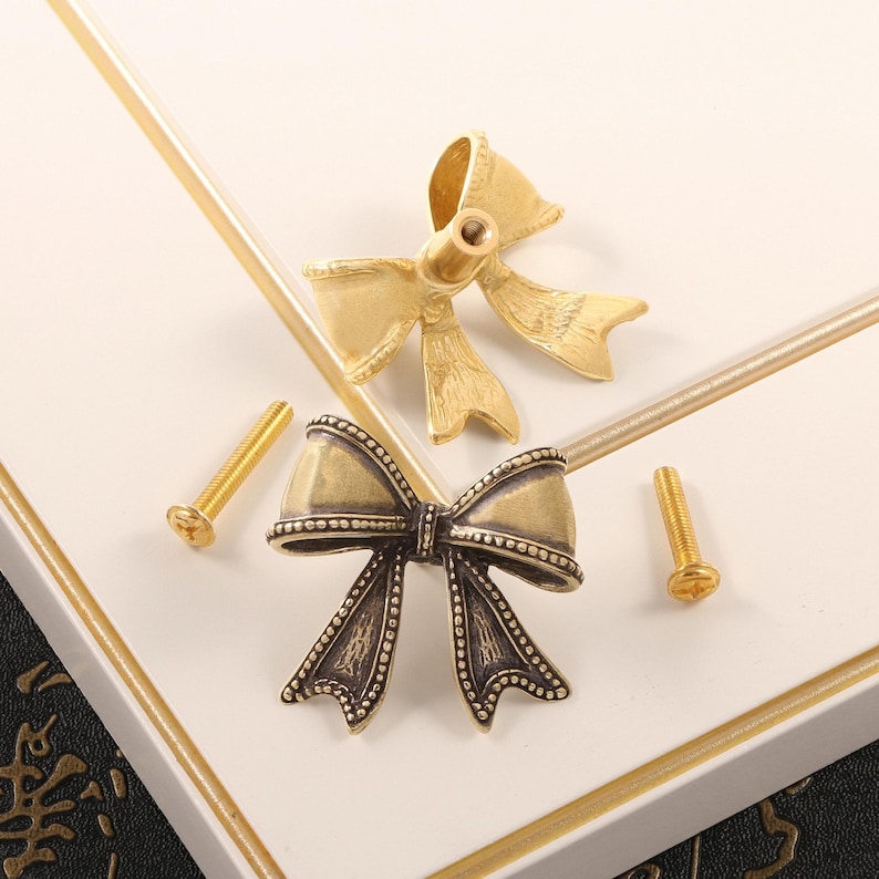 May include: Two gold-coloured bow-shaped cabinet pulls with decorative detailing, and two matching screws. The pulls are set against a white surface with gold trim, suggesting a furniture hardware theme.