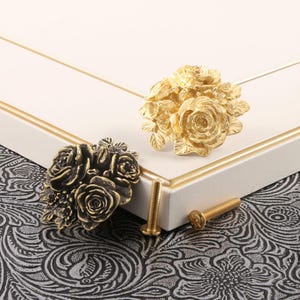 Könnte beinhalten: Zwei dekorative Schrankknöpfe mit einem Rosenstrauß-Design. Einer ist goldfarben, der andere bronzefarben. Zwei goldfarbene Schrauben sind ebenfalls sichtbar. Die Knöpfe befinden sich auf einer weißen Schranktür mit goldenem Rand, vor einem gemusterten Hintergrund.