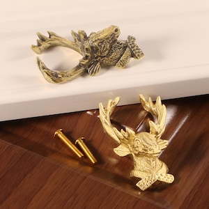 Può includere: Due pomelli per mobili color ottone a forma di testa di cervo e corna. I pomelli sono rifiniti con una finitura vintage. Sono visibili anche due viti in ottone. I pomelli sono su una superficie di legno.