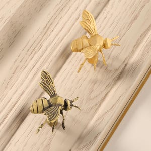 Könnte beinhalten: Zwei dekorative Möbelgriffe in Bienenform. Die Griffe sind aus goldfarbenem Metall gefertigt und weisen aufwendige Details wie Flügel und Fühler auf. Die Bienen sind auf einer hellen Holzoberfläche positioniert.