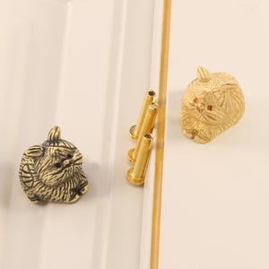 Könnte beinhalten: Zwei messingfarbene Schrankknöpfe in Katzenform mit detaillierter Fellstruktur. Drei goldene Schrauben sind ebenfalls sichtbar. Die Knöpfe befinden sich auf einer weißen Oberfläche mit goldenem Akzent.