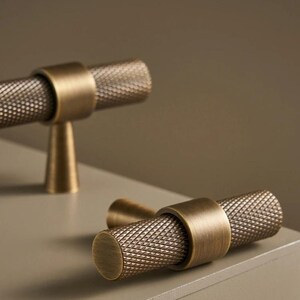 May include: Close-up of two antique brass cabinet pulls. The pulls have a cylindrical, textured handle with a diamond pattern and are attached to a round base. The word "NURLIN" is printed on a blue rectangle in the background.